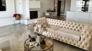 A modern living room with a beige tufted sofa, a glass coffee table with a laptop, a fireplace, and a kitchen area in the background. - LUXURY WELLNESS PENTHOUSE KADENA WITH DREAM SEE VIEW (Zadar)