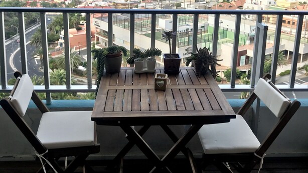 A balcony with a wooden table and two white chairs, overlooking a cityscape with buildings and a port. - Central and large apartment with sea views (Las Palmas de Gran Canaria)