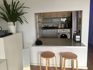Uma cozinha compacta com máquina de lavar, torradeira e pia. Há dois bancos em frente ao balcão.