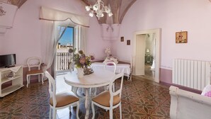 A room with a white round table, white chairs, a television on a shelf, and a balcony with a view.
