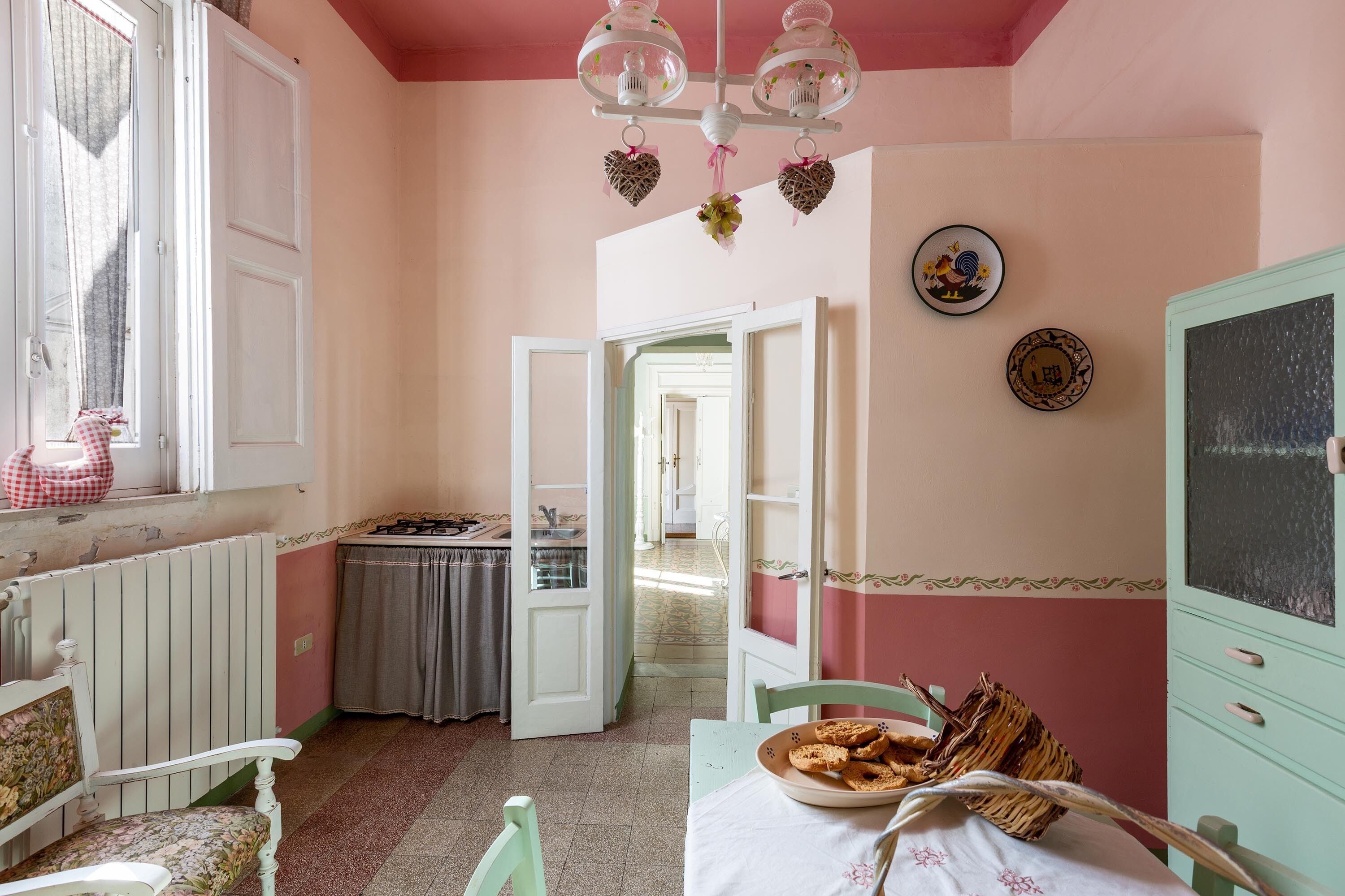 A room with a dining table, chairs, a radiator, a window with curtains, a door leading to another room, and a wall with decorative plates.