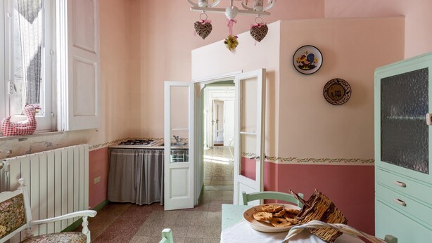 A room with a dining table, chairs, a radiator, a window with curtains, a door leading to another room, and a wall with decorative plates. - 3267 Casa Romantica by Barbarhouse (Sava)