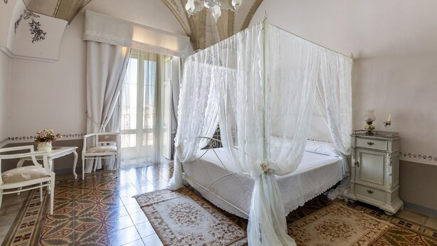 A bedroom with a four-poster bed, a nightstand, a chair, and a table with a vase of flowers.