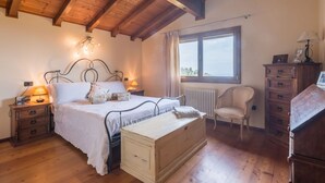 Ein Schlafzimmer mit Holzbalkendecke, einem Bett mit weißer Bettwäsche, einem Nachttisch mit Lampe, einem Schminktisch und einem Stuhl.