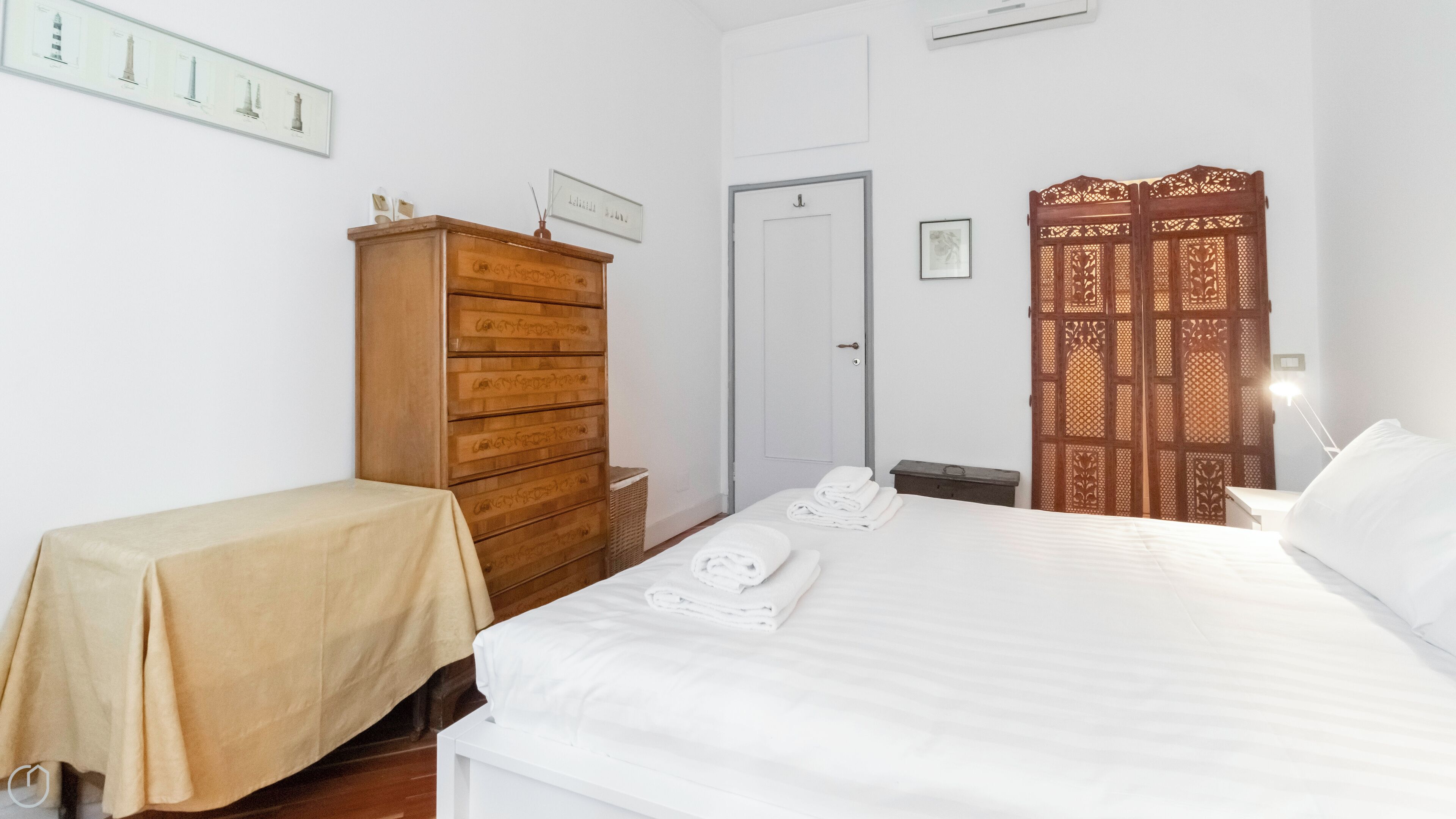 A hotel room with a bed, a wooden dresser, a small table with a tablecloth, a door, and a decorative screen.