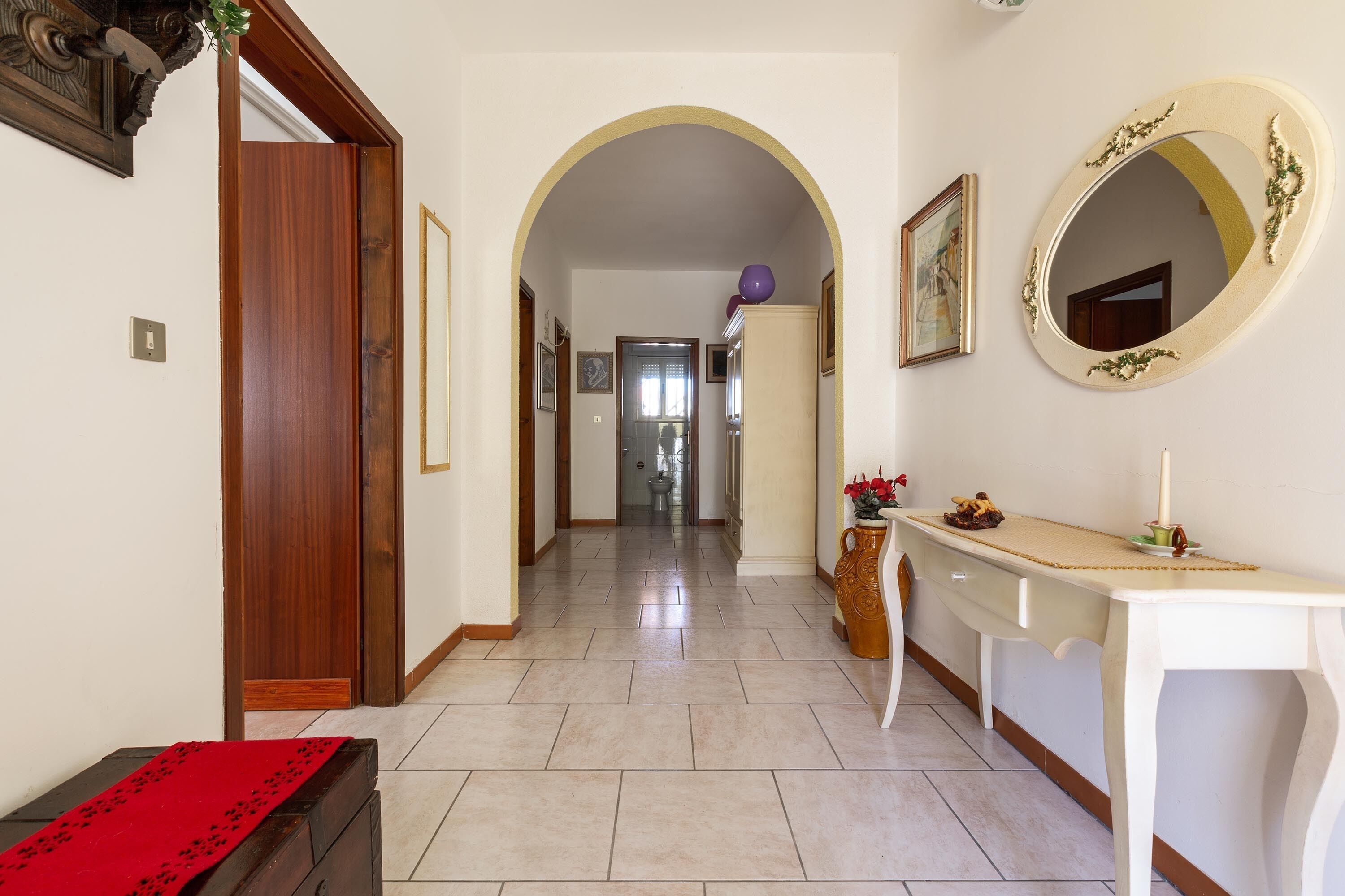Un corridoio con una porta in legno, un tavolino consolle bianco con uno specchio e un tappeto rosso su un settimino.