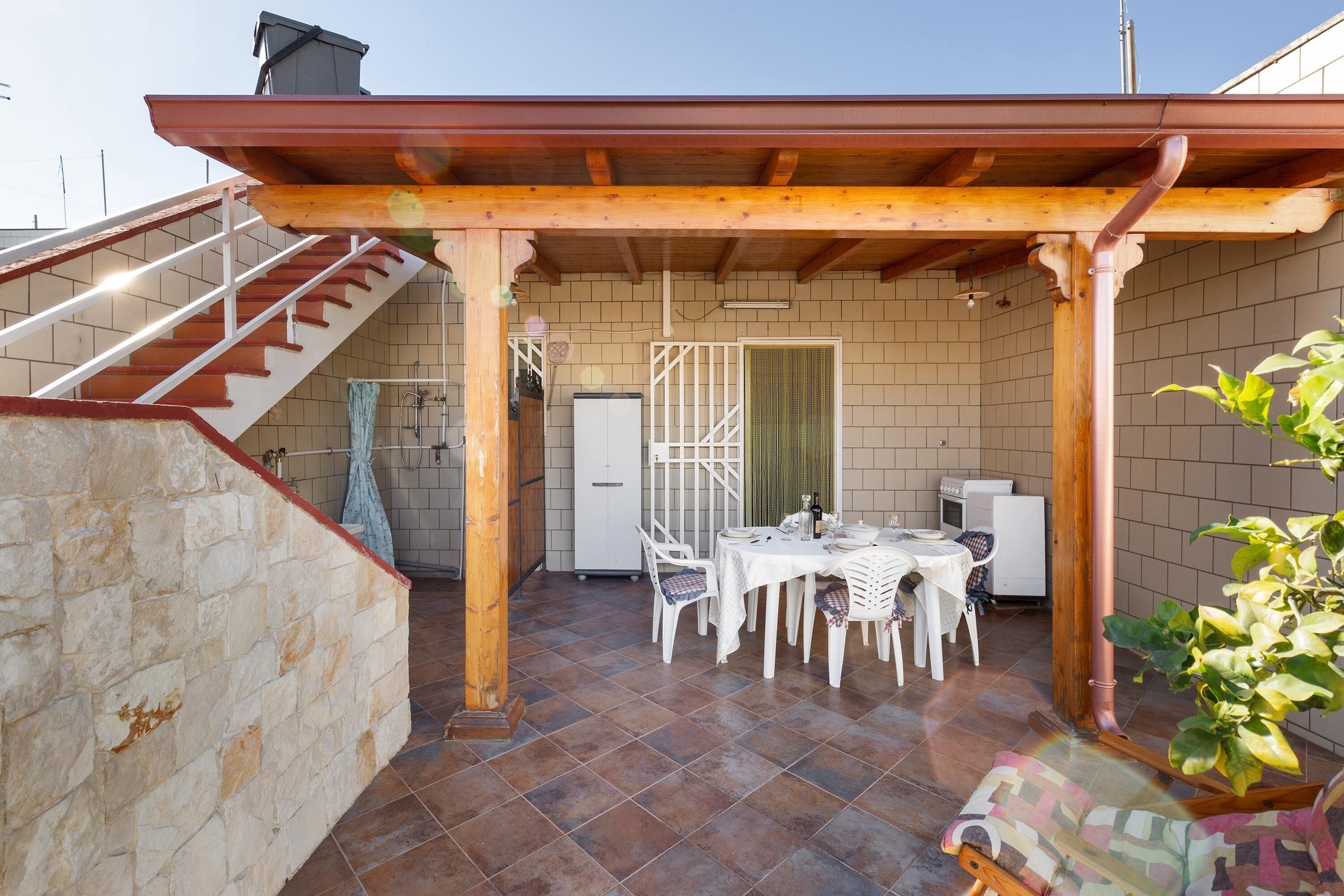 Eine überdachte Terrasse mit Esstisch und Stühlen, einer Treppe und einem Kühlschrank.