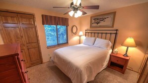 Une chambre à coucher comprenant un lit, un ventilateur de plafond, une fenêtre avec des stores, une table de chevet avec une lampe et une commode en bois.