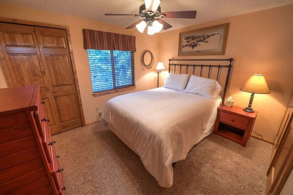 Une chambre à coucher comprenant un lit, un ventilateur de plafond, une fenêtre avec des stores, une table de chevet avec une lampe et une commode en bois.