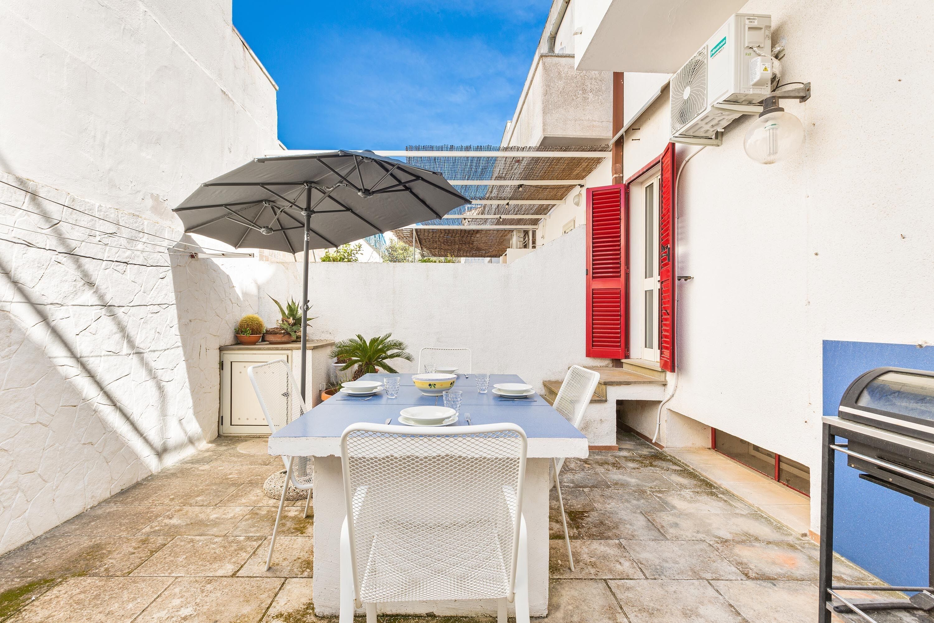 A patio with a table set for two, a white umbrella, and a red shuttered door.