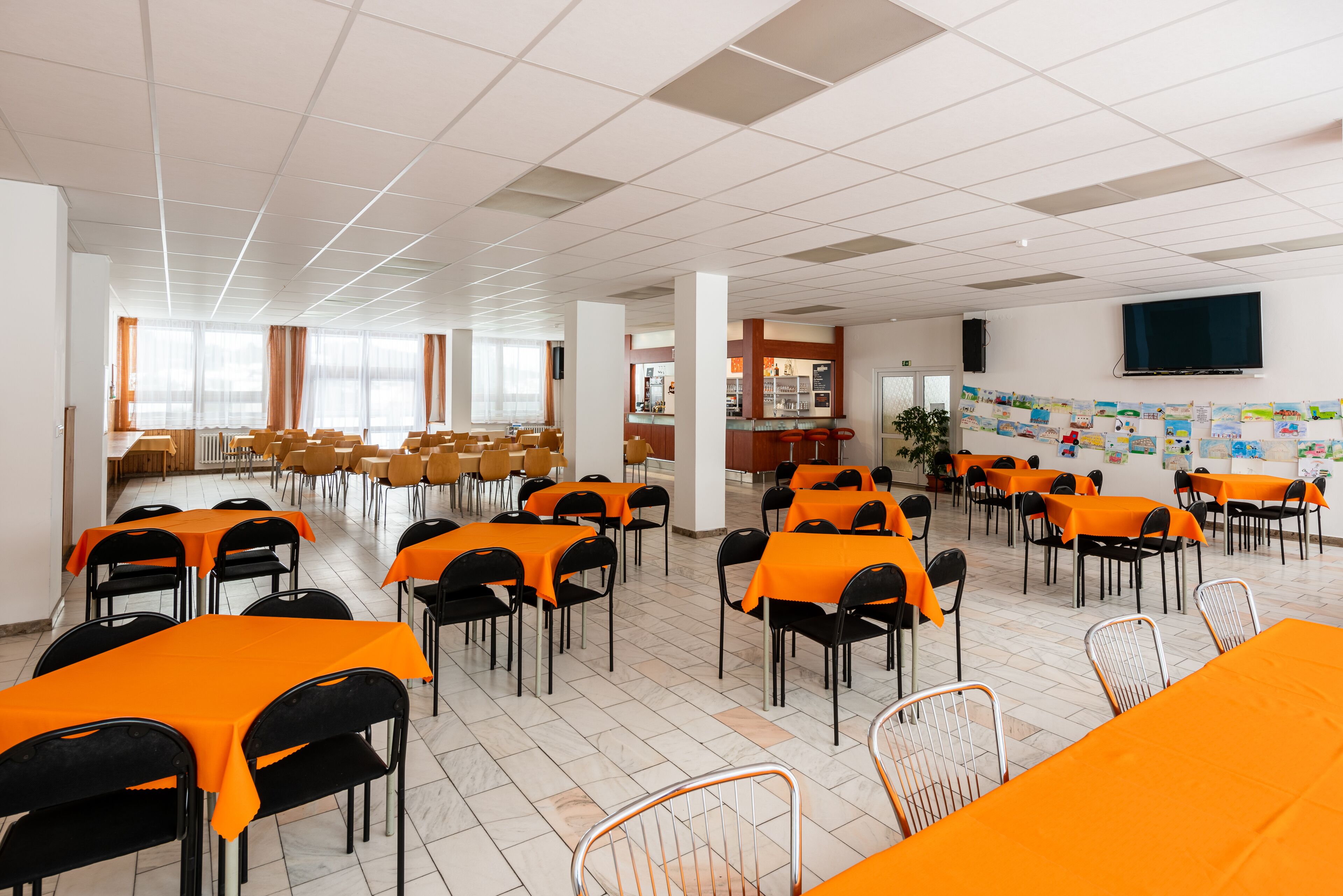 A spacious dining area with tables covered in orange tablecloths and black chairs, featuring a tiled floor and a large window with curtains.