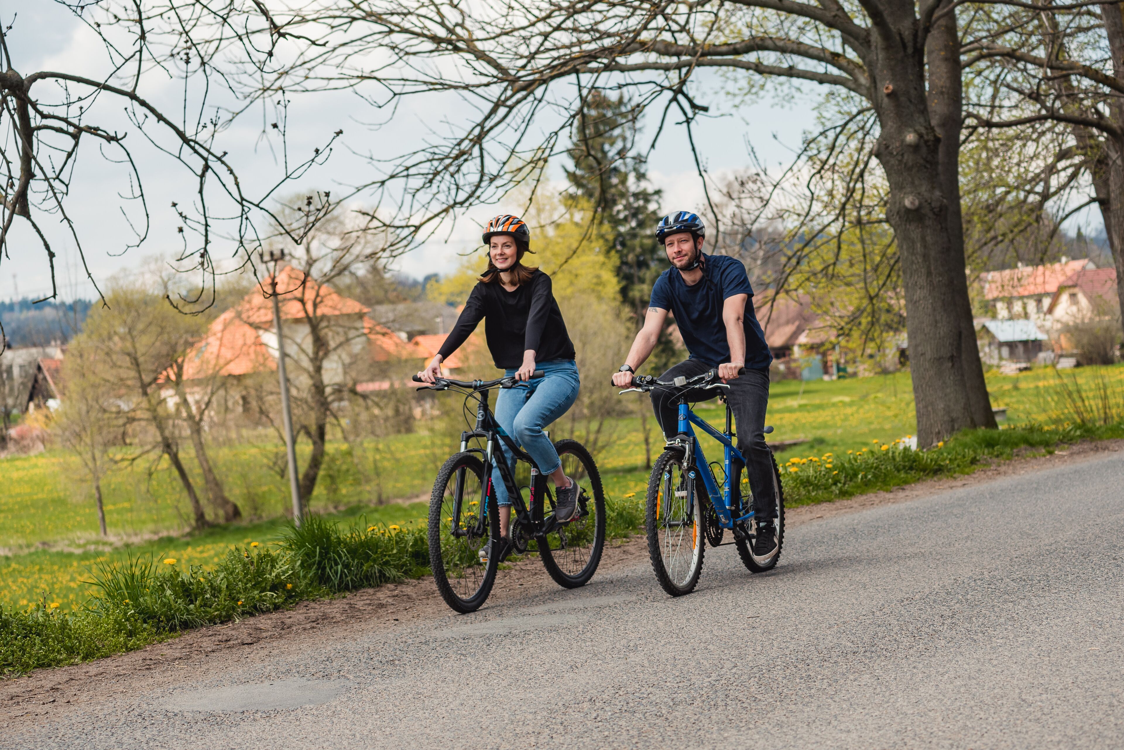 Två personer cyklar på en asfalterad stig, med träd och hus i bakgrunden.