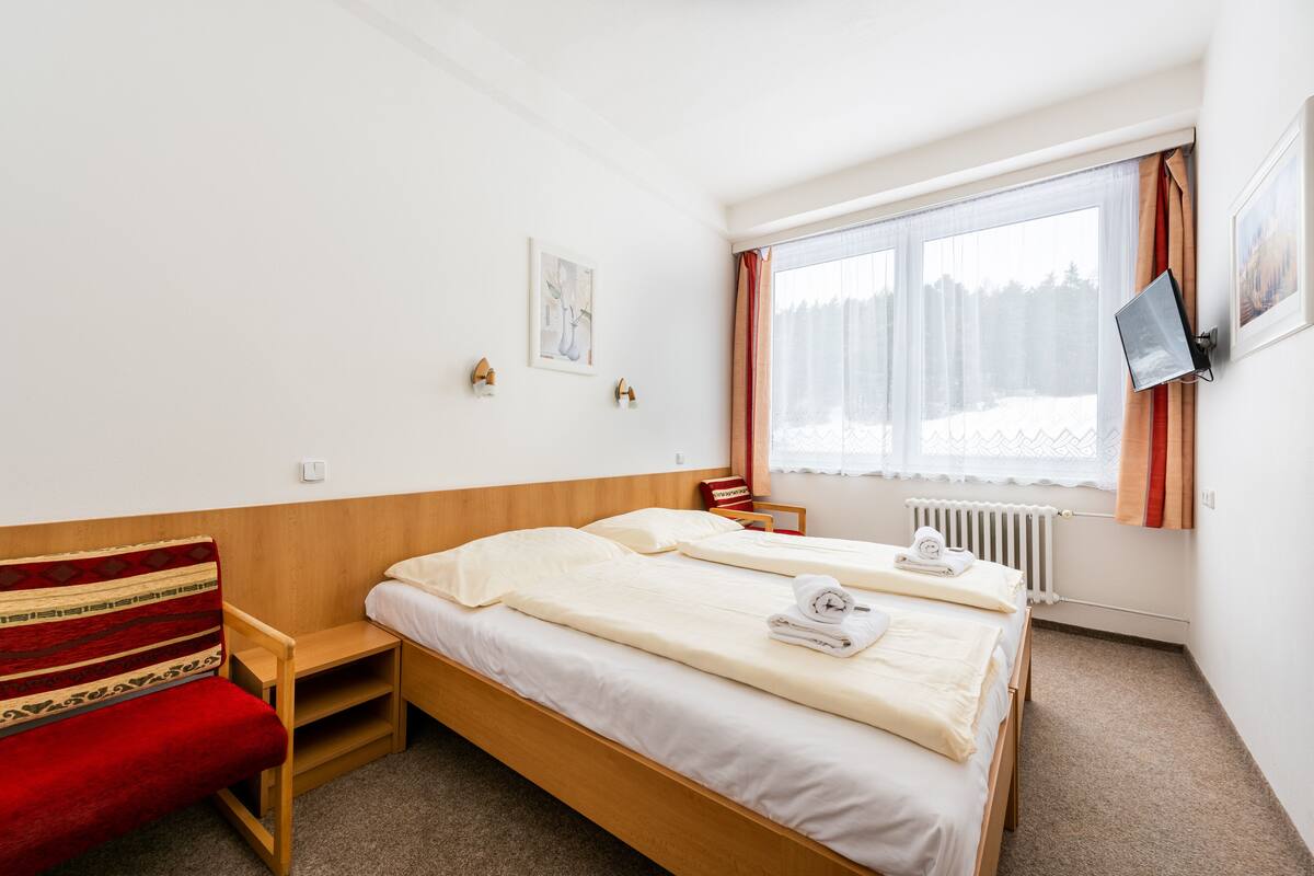 A hotel room with a wooden bed, a red sofa, a small table, a window with curtains, and a wall-mounted TV.
