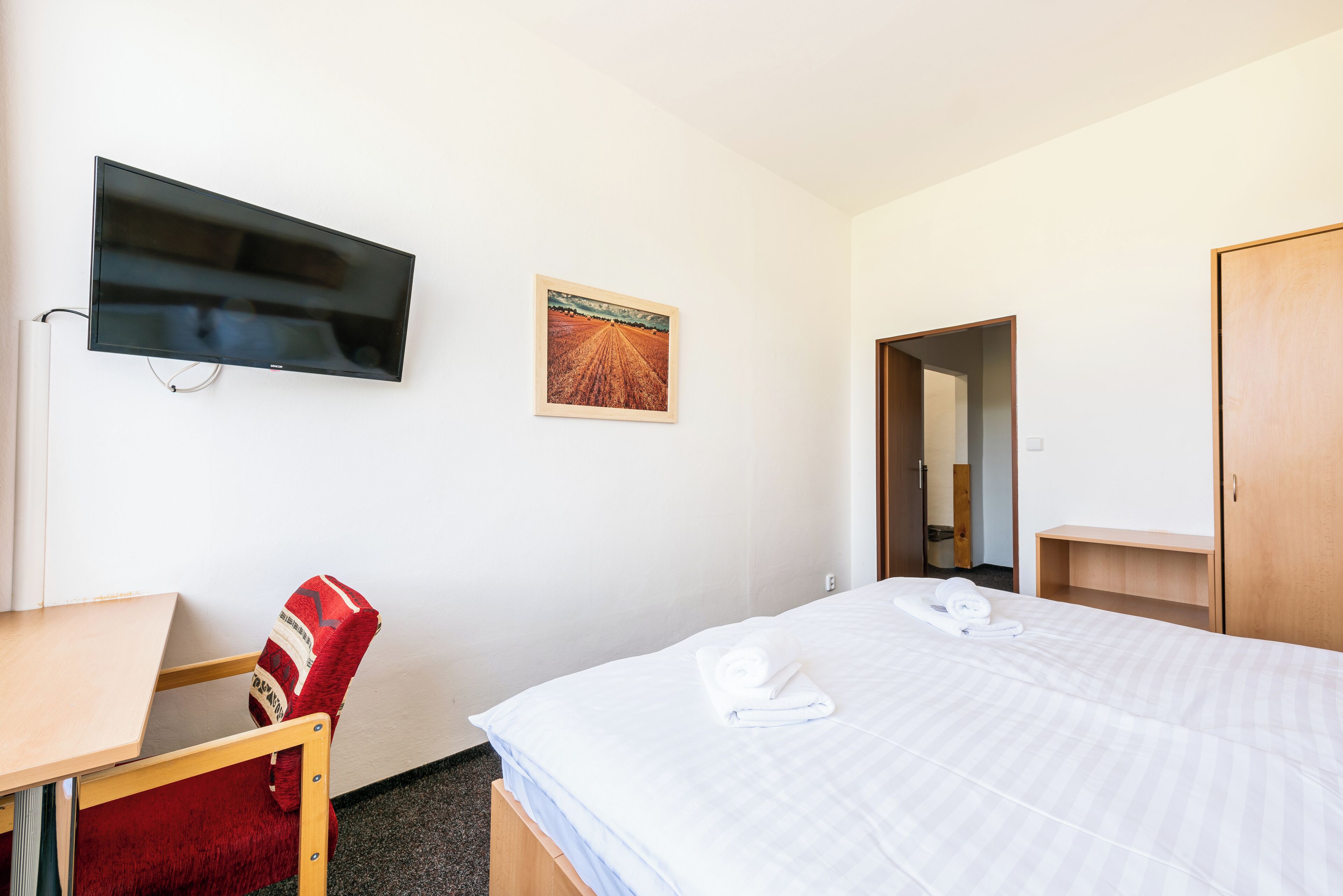 A hotel room with a bed, a TV mounted on the wall, a painting, a wooden chair, and a wooden wardrobe.