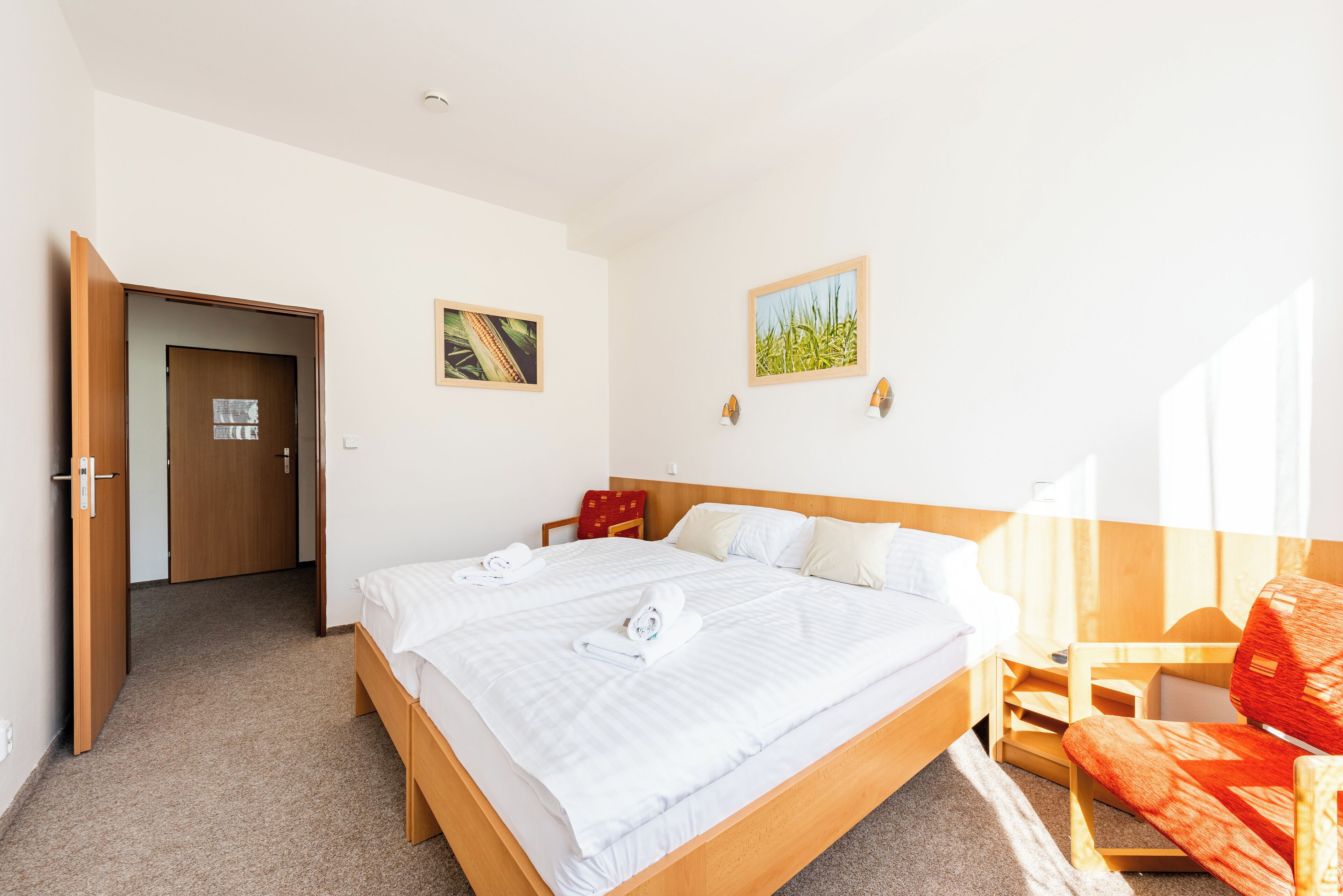 Habitación de hotel con una cama grande, cabecera de madera, una silla roja y dos cuadros en la pared.