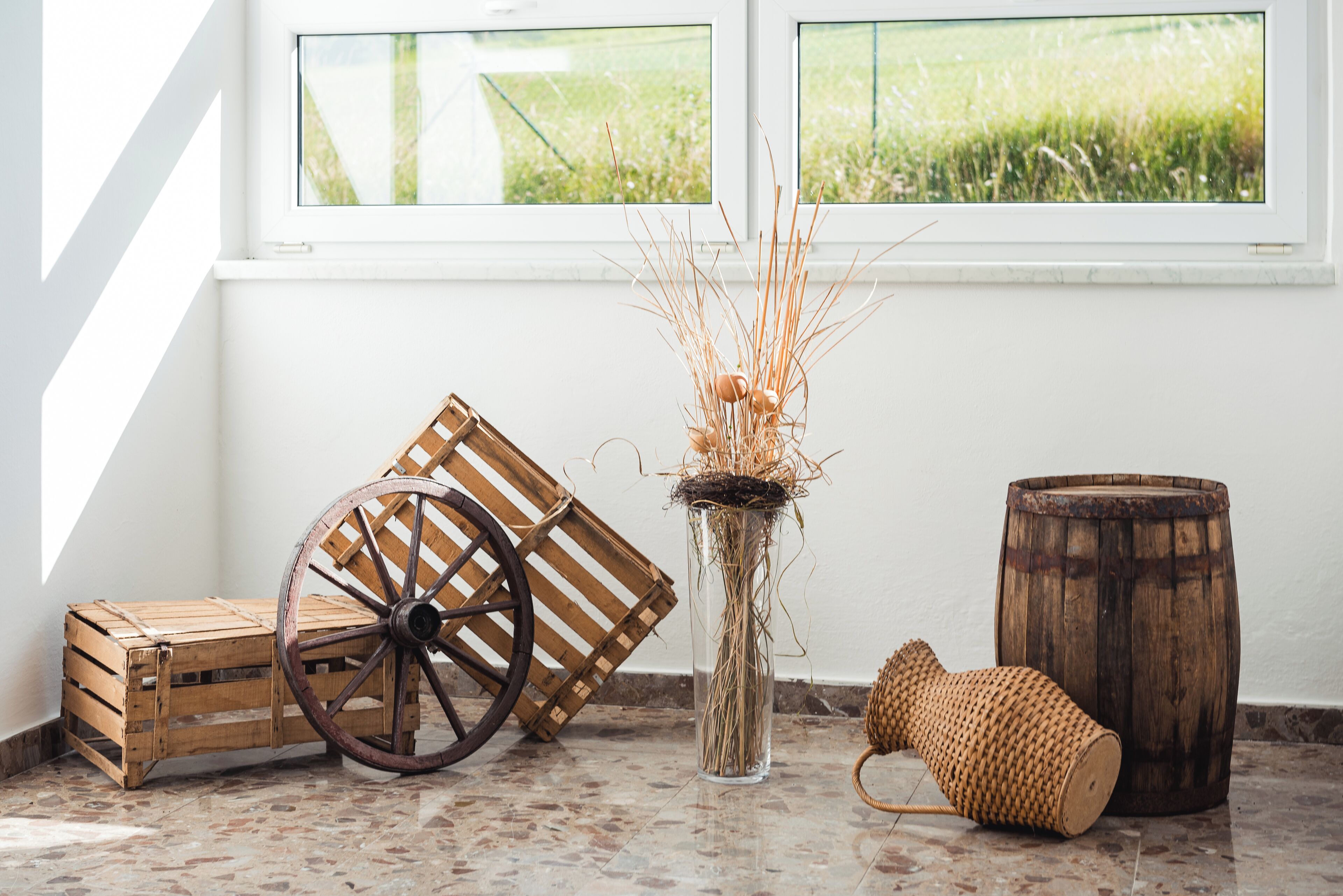 Un intérieur rustique agrémenté d’une caisse en bois, d’une roue de charrette ancienne, d’un vase contenant des plantes séchées, d’un panier tressé et d’un tonneau en bois.