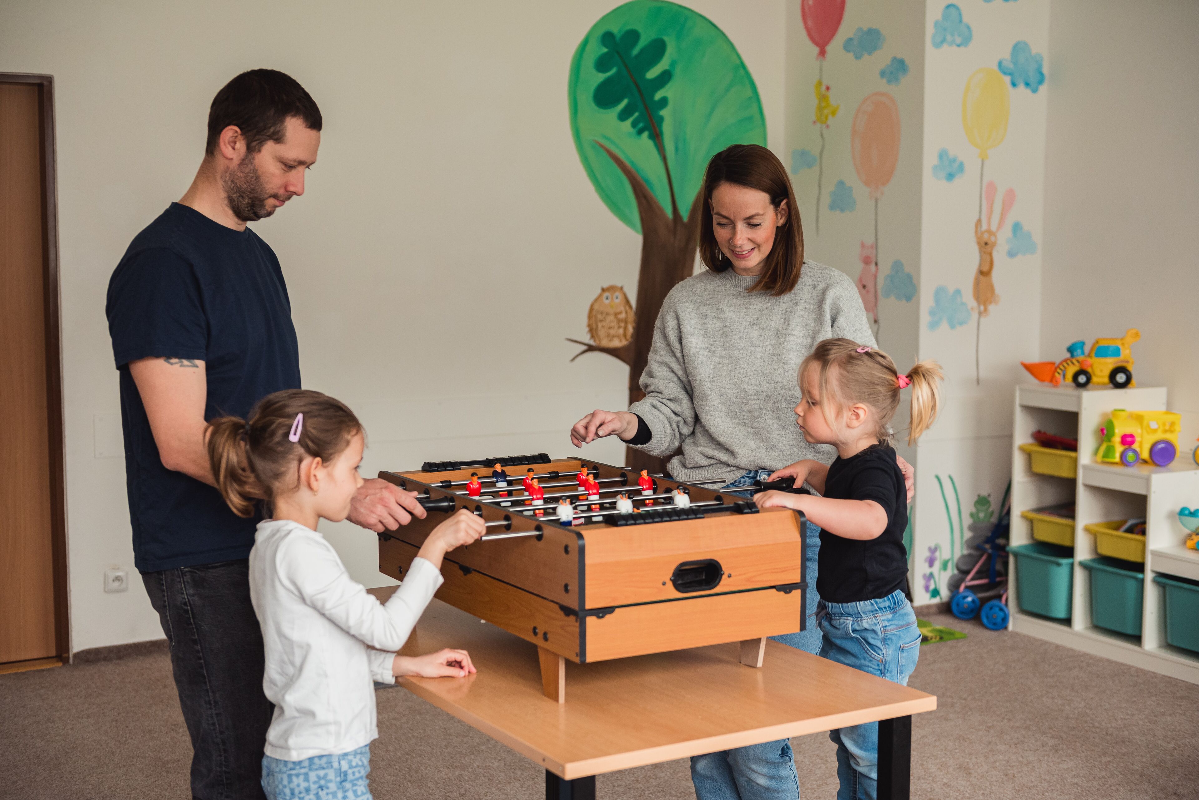 A family playing foosball in a room with a wooden table, colorful wall decorations, and storage bins in the background.