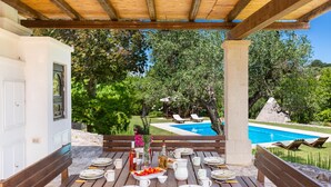 Un espace de restauration en plein air avec une table en bois dressée pour un repas, offrant une vue sur une piscine et une végétation luxuriante.