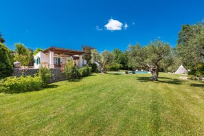 A well-maintained lawn with a stone wall, a white building with green shutters, and a tree.