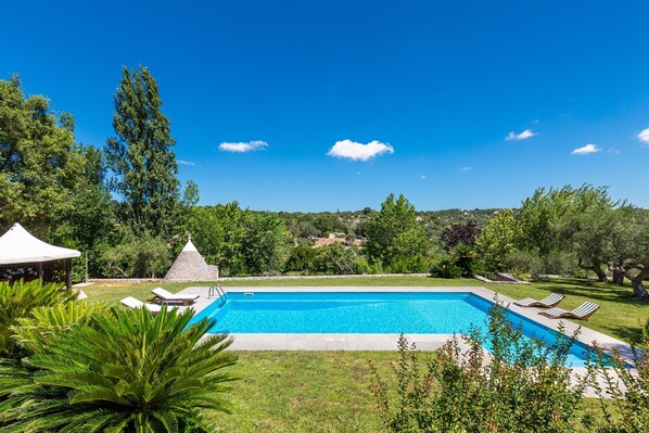 A rectangular swimming pool with lounge chairs and a gazebo in a lush, green landscape.