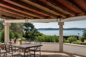 Un espace extérieur abrité, avec une table et des chaises, offrant une vue sur un plan d’eau et des collines.