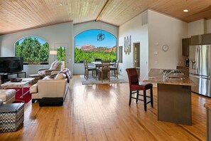 A spacious living area with a dining table, seating, a kitchen with a stainless steel refrigerator, and large windows with a view of greenery and a building.