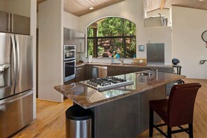 A modern kitchen with stainless steel appliances, a large island with a gas stove, and a view of the outdoors through a large window.