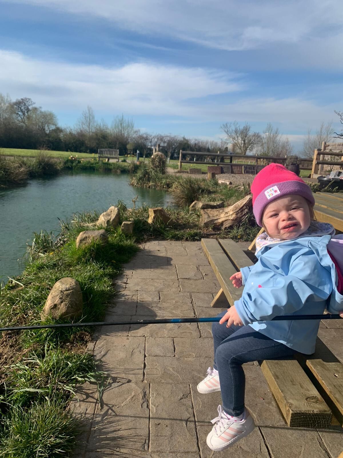 A child sitting on a wooden bench by a pond with a clear sky in the background.