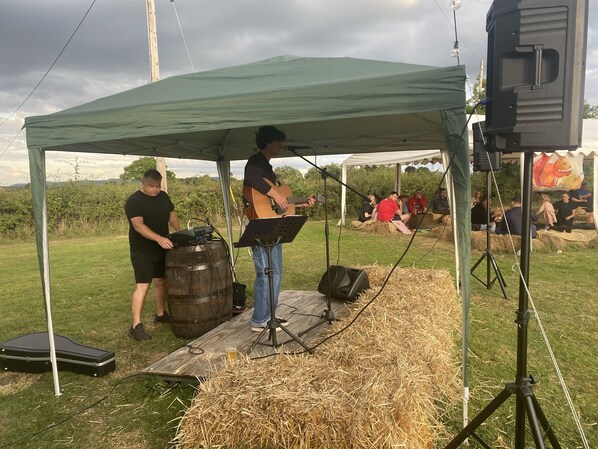 Un spectacle de musique en plein air, sous une tente, avec une scène faite de bottes de foin, une grande enceinte et un guitariste.