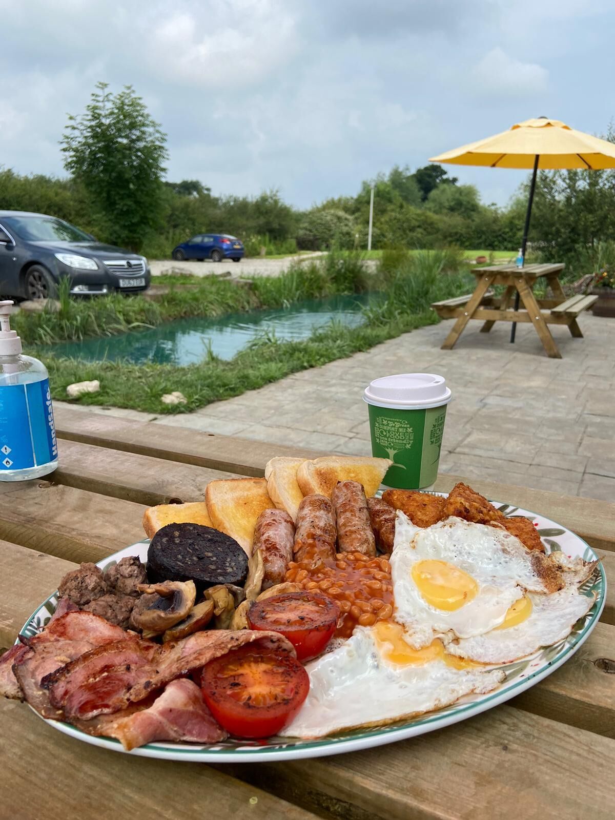 A breakfast plate with eggs, sausages, beans, bacon, and toast on a wooden table outdoors.