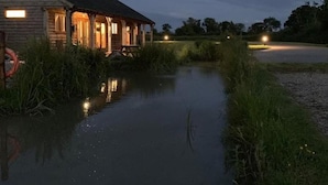 A rustic wooden building with a porch, a small waterway in front, and a road with a vehicle in the distance. - Beech Pod Cabin in Beautiful Surroundings Wrexham (Wrexham)