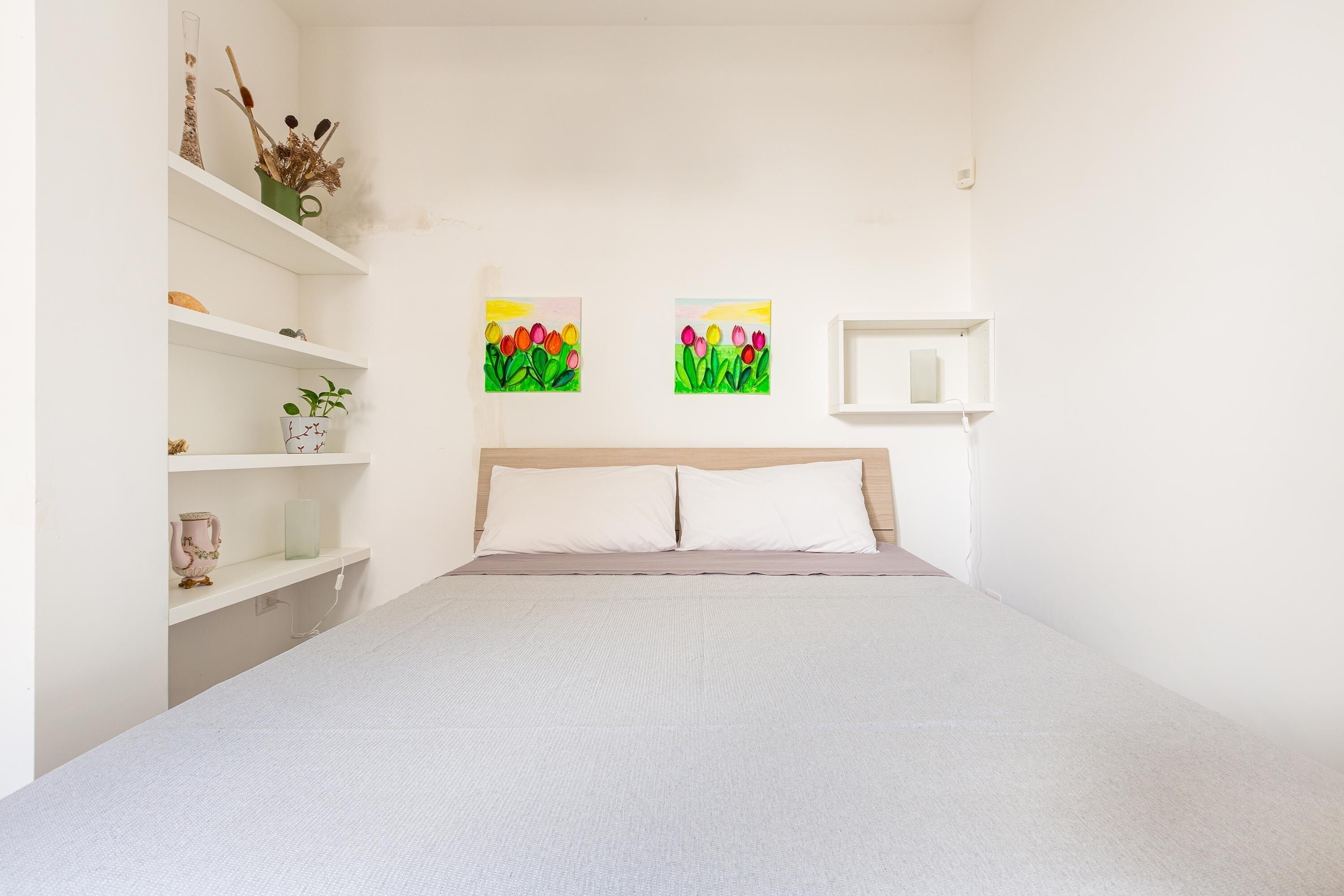 Un dormitorio con cama, paredes blancas y dos cuadros de flores.