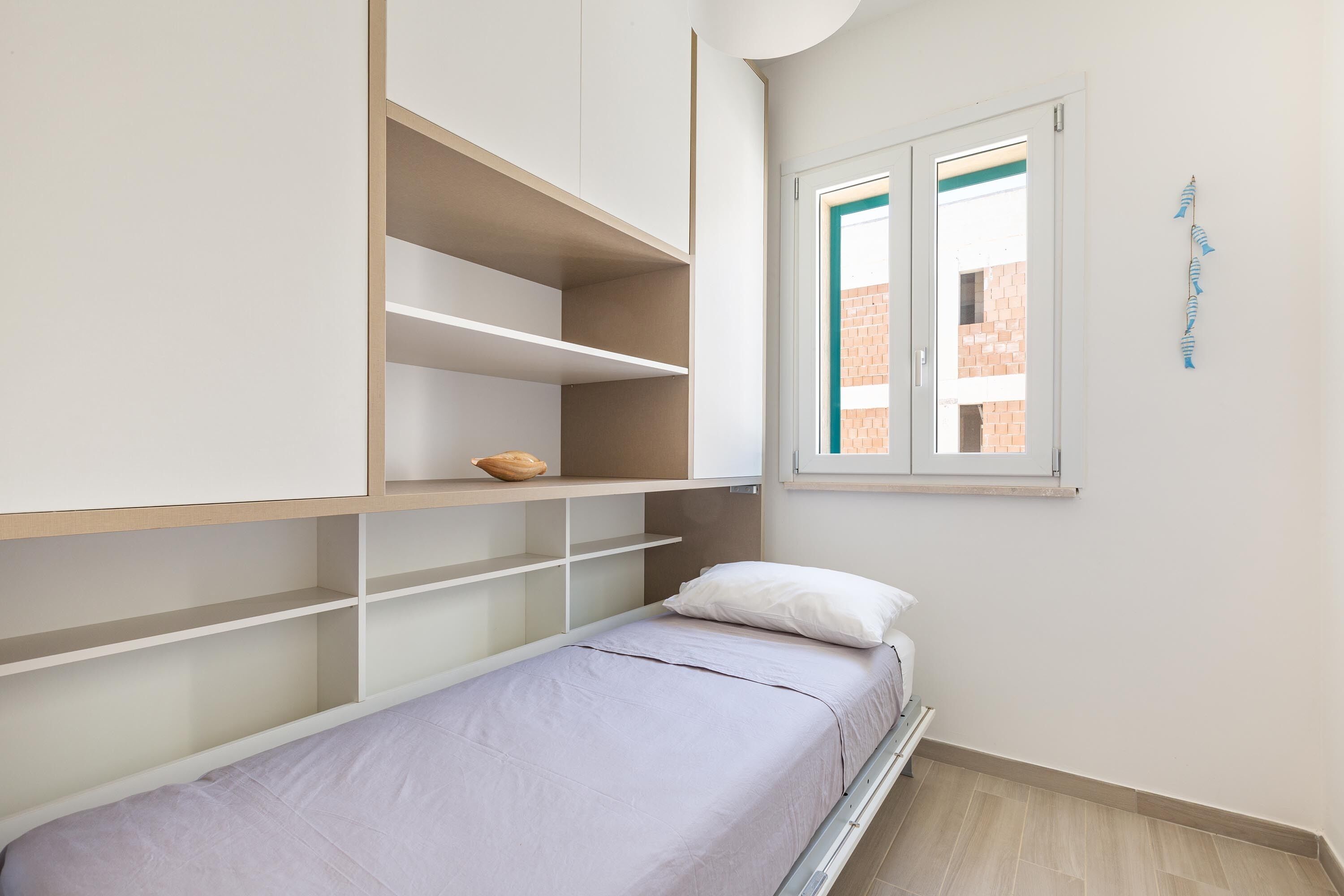 A small, neatly organized room with a bed, built-in shelving, and a window with a view of a brick building.