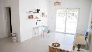 A bright, modern living room with a white sofa, a coffee table, and a lamp. There is a desk with a chair and a plant on the wall. - Villa Ravanica (Giannuzza) with pool 400m from the beach (Marina di Ragusa)