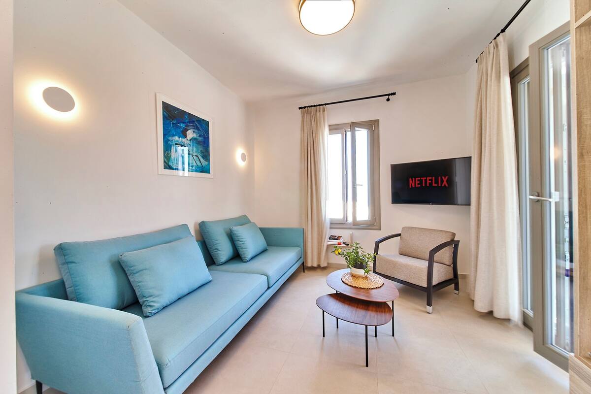 A modern living room with a light blue sofa, a round wooden coffee table, a beige armchair, a flat-screen TV mounted on the wall, and a window with curtains.