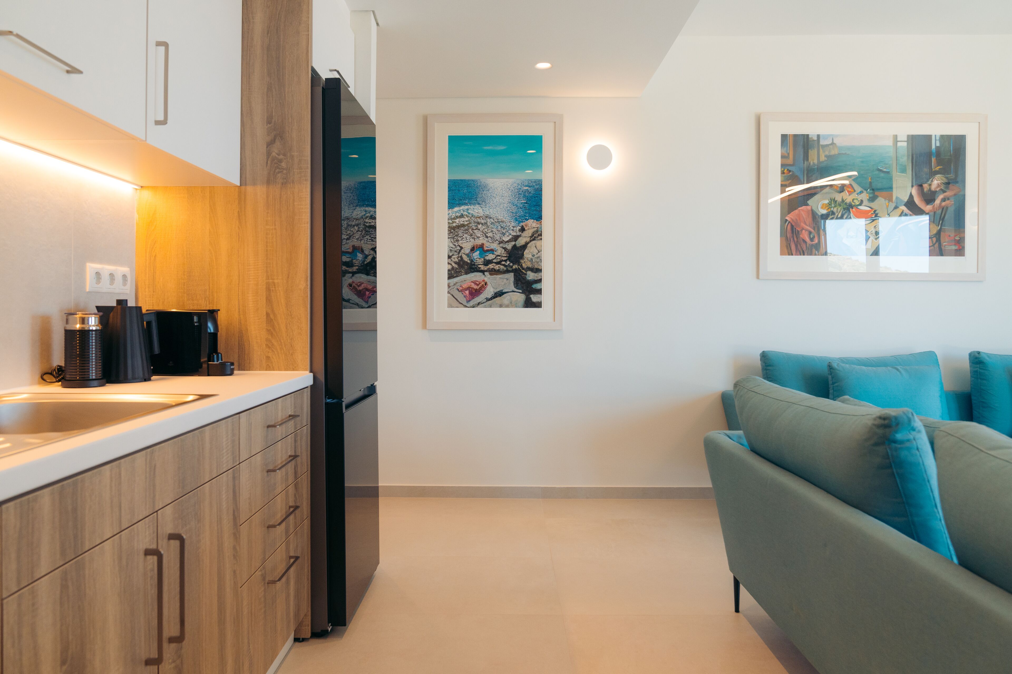 A modern kitchen with wooden cabinets, a black refrigerator, and a coffee maker on the counter. There are two framed artworks on the wall, one depicting a rocky landscape and the other a beach scene. A teal sofa is positioned in the living area.