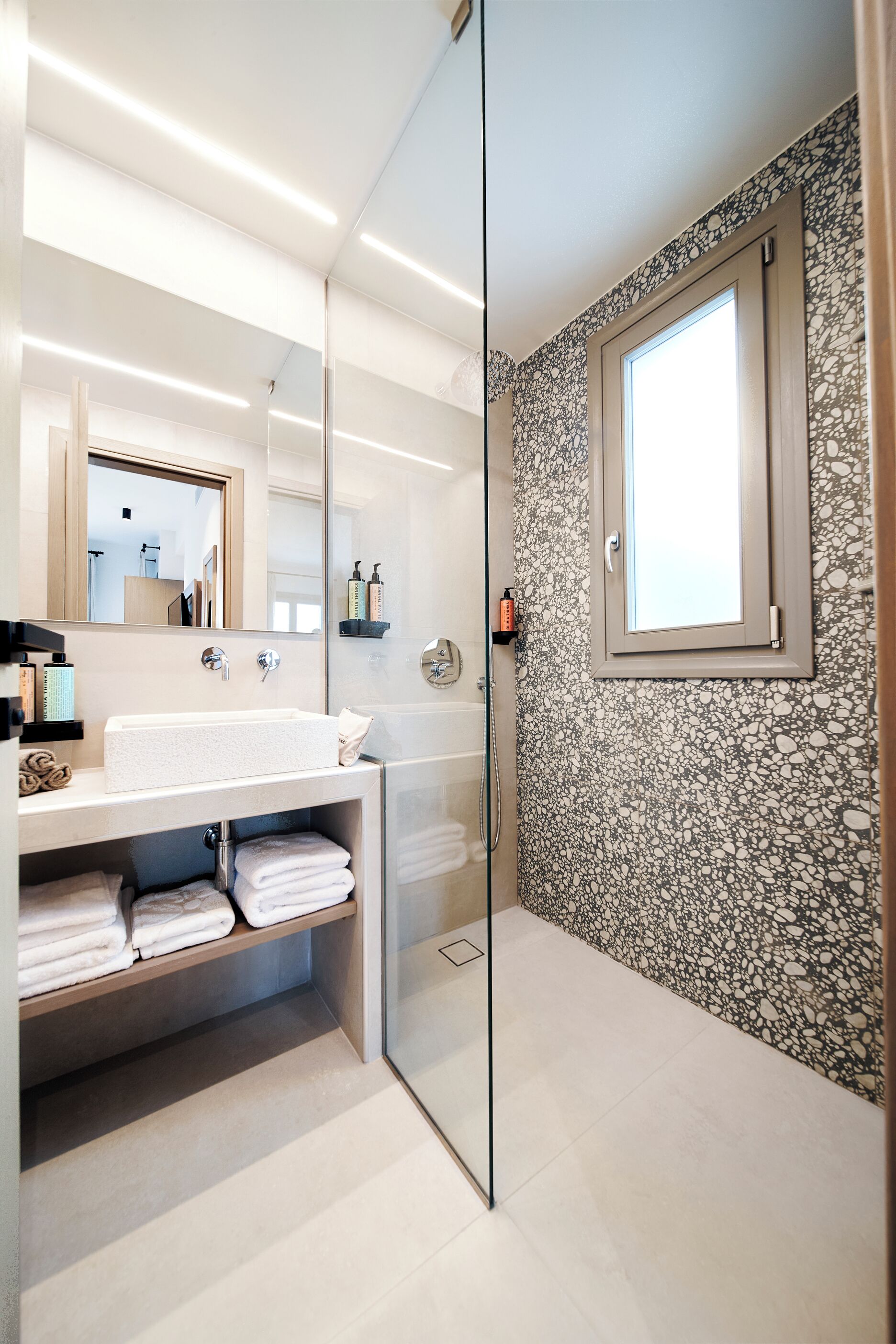 Un baño moderno con cabina de ducha de vidrio, un espejo grande, un lavabo blanco y una pared con diseño de piedra.
