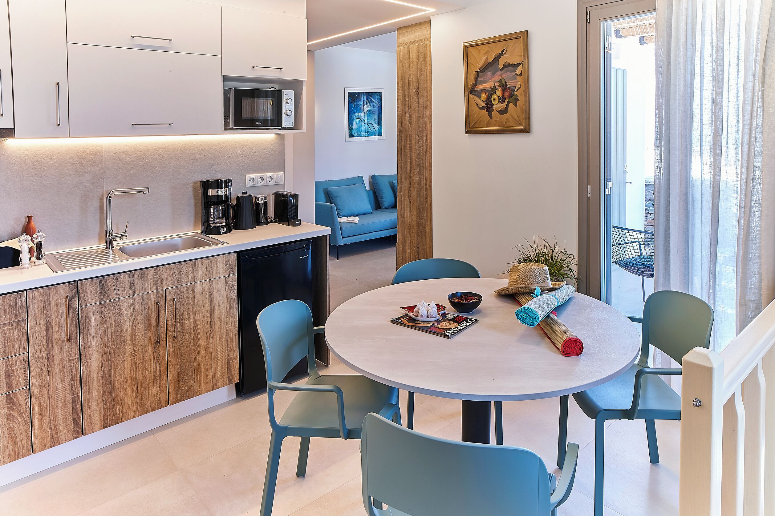 A modern kitchen with a sink, microwave, and coffee maker. There is a dining area with a round table and chairs. A sofa and a painting are visible in the background.
