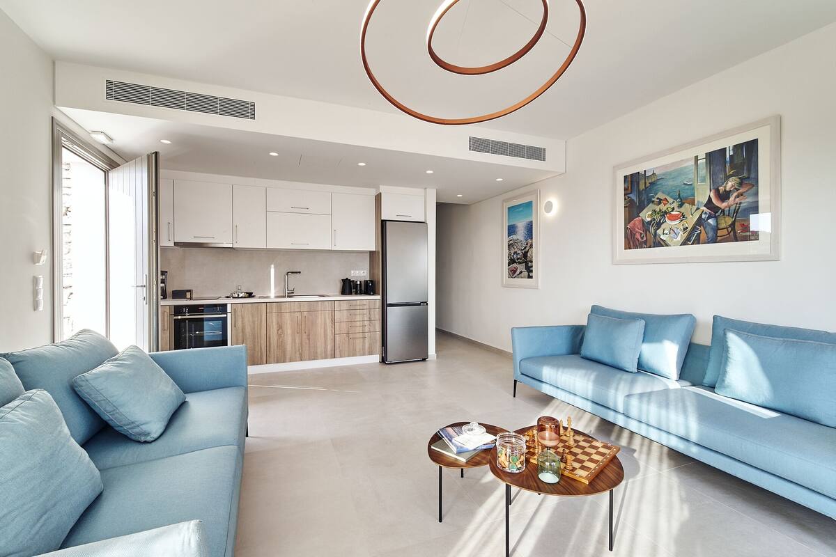 A modern living room with a blue sofa, a coffee table with a chess set, and a kitchen in the background.