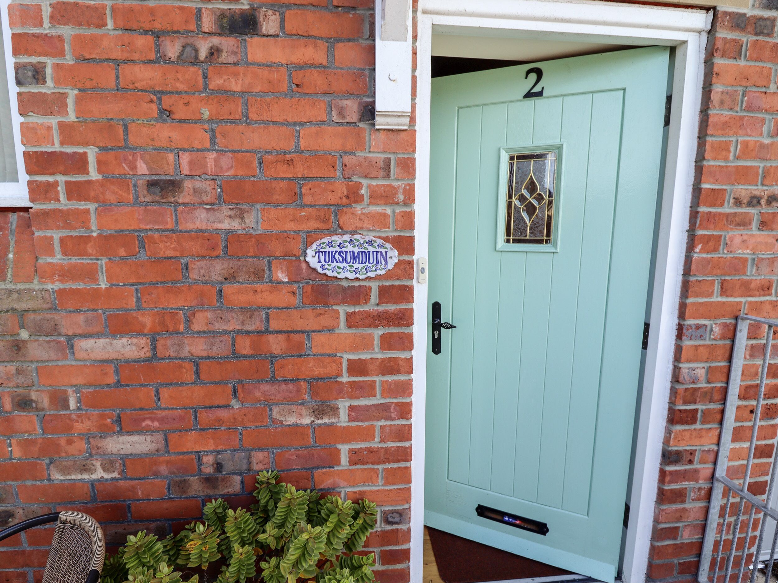 Una porta verde chiaro con il numero '2' sopra, una piccola finestra con un motivo decorativo in vetro e un cartello sulla parete in mattoni che recita 'TUKSUMDUIN'.