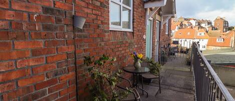 A brick building with a balcony featuring a table and chairs, a potted plant, and a decorative fence.
