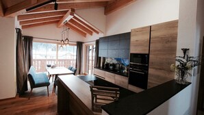 Una cocina moderna con vigas de madera, una zona de comedor con sillas azules y vistas a una terraza de madera.