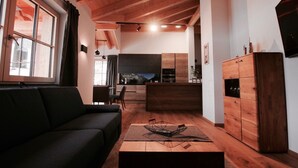 Una sala de estar moderna con un sofá oscuro, piso de madera y una cocina al fondo.