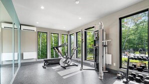 Une salle de sport moderne, équipée de matériel d’exercice, dotée de grandes fenêtres et offrant une vue sur les arbres.