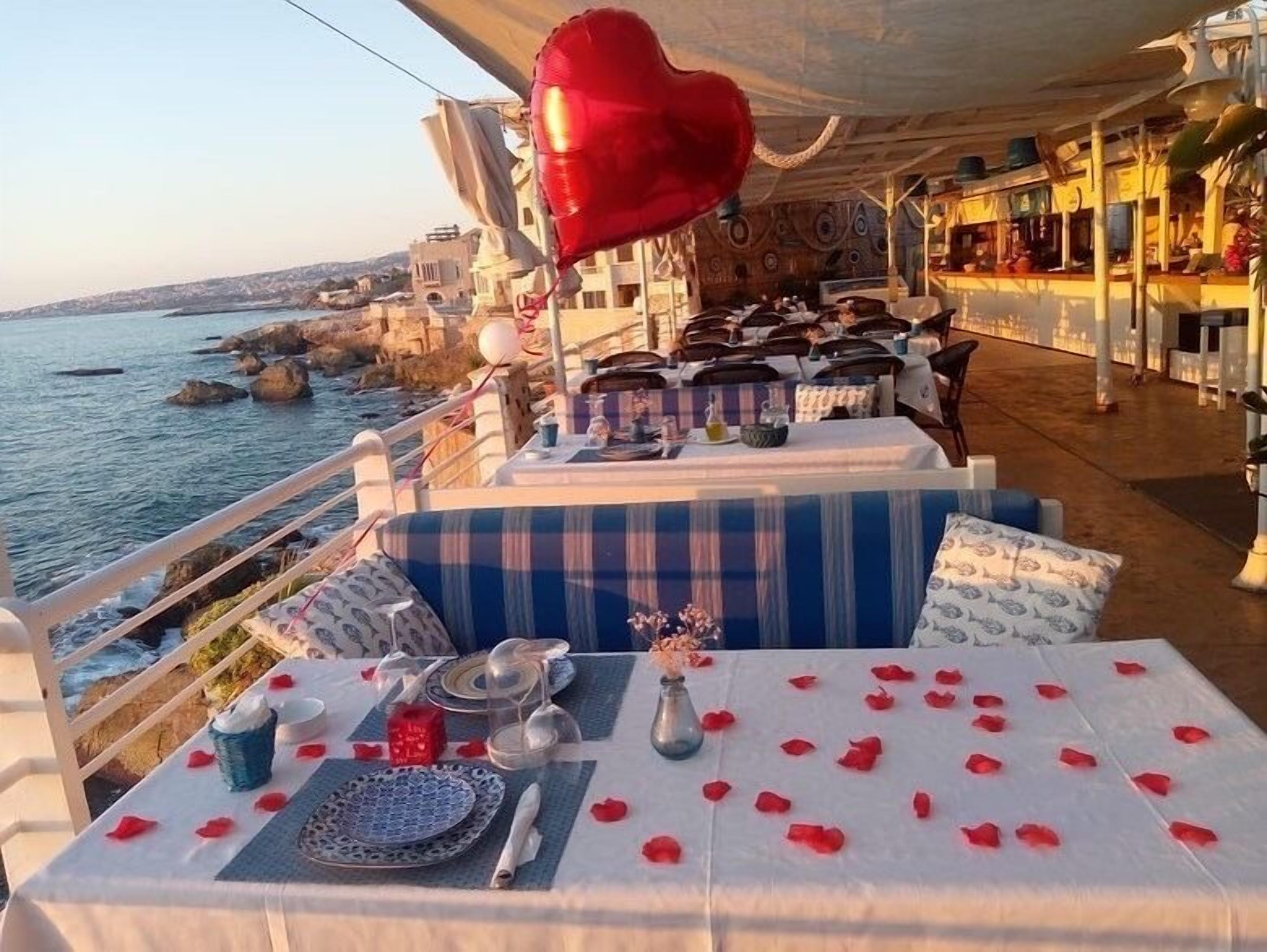 Un restaurante frente al mar con una mesa para dos, decorada con un globo con forma de corazón, pétalos de rosa y vistas al mar y a la costa rocosa.