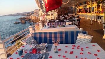 Un restaurante frente al mar con una mesa para dos, decorada con un globo con forma de corazón, pétalos de rosa y vistas al mar y a la costa rocosa.