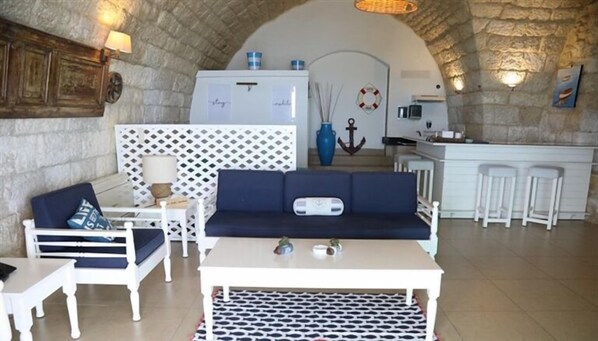 A cozy living area with a sofa, a small table, and a white coffee table. There is a kitchen area in the background with a refrigerator and bar stools.