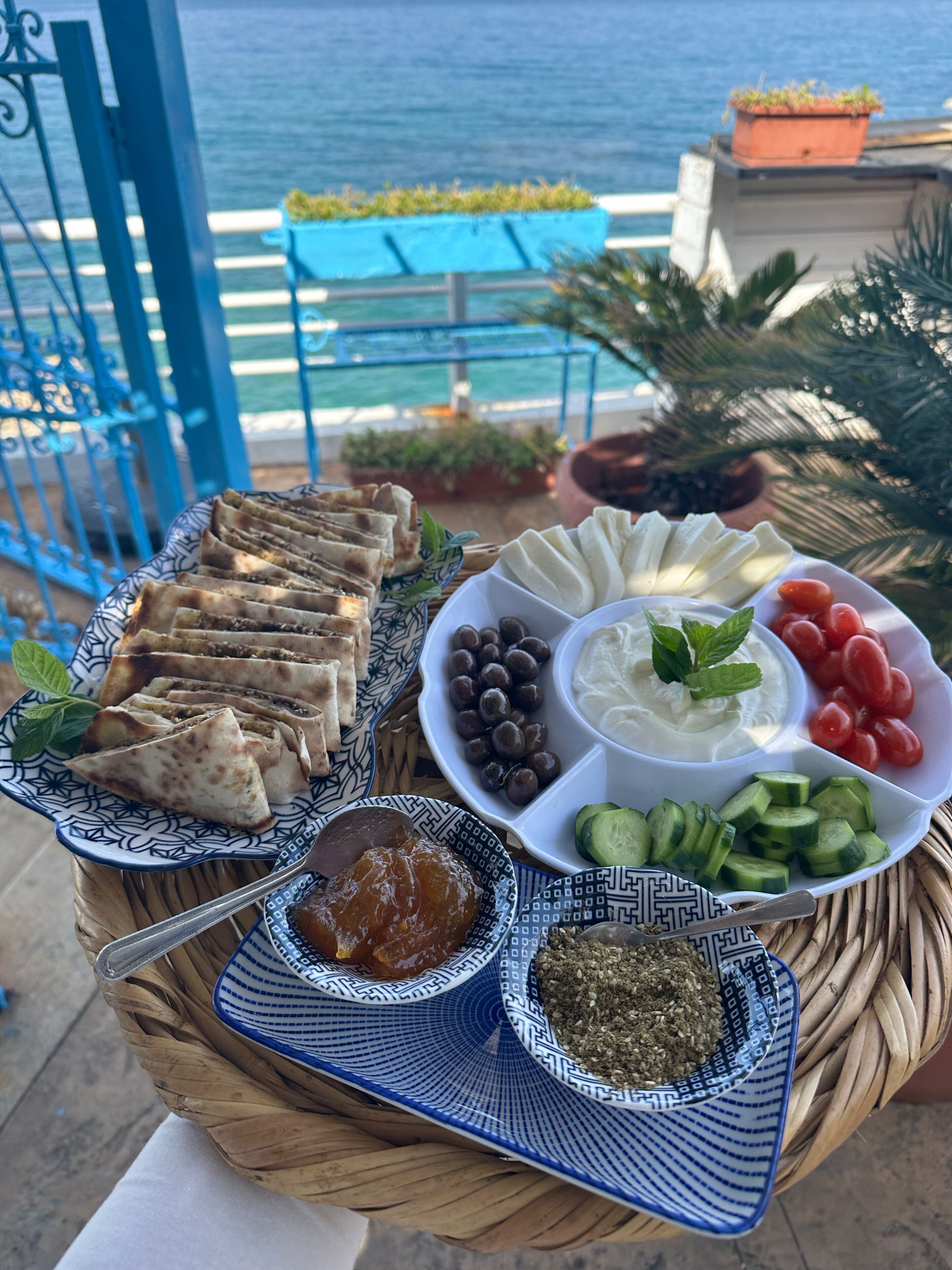 A tray of food with various dishes including flatbreads, olives, tomatoes, cucumbers, and dips, set on a balcony with a view of the sea.