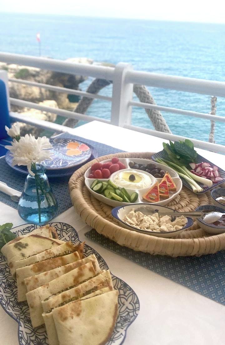 A table with a variety of food items including flatbreads, hummus, olives, and vegetables, set against a scenic ocean view.