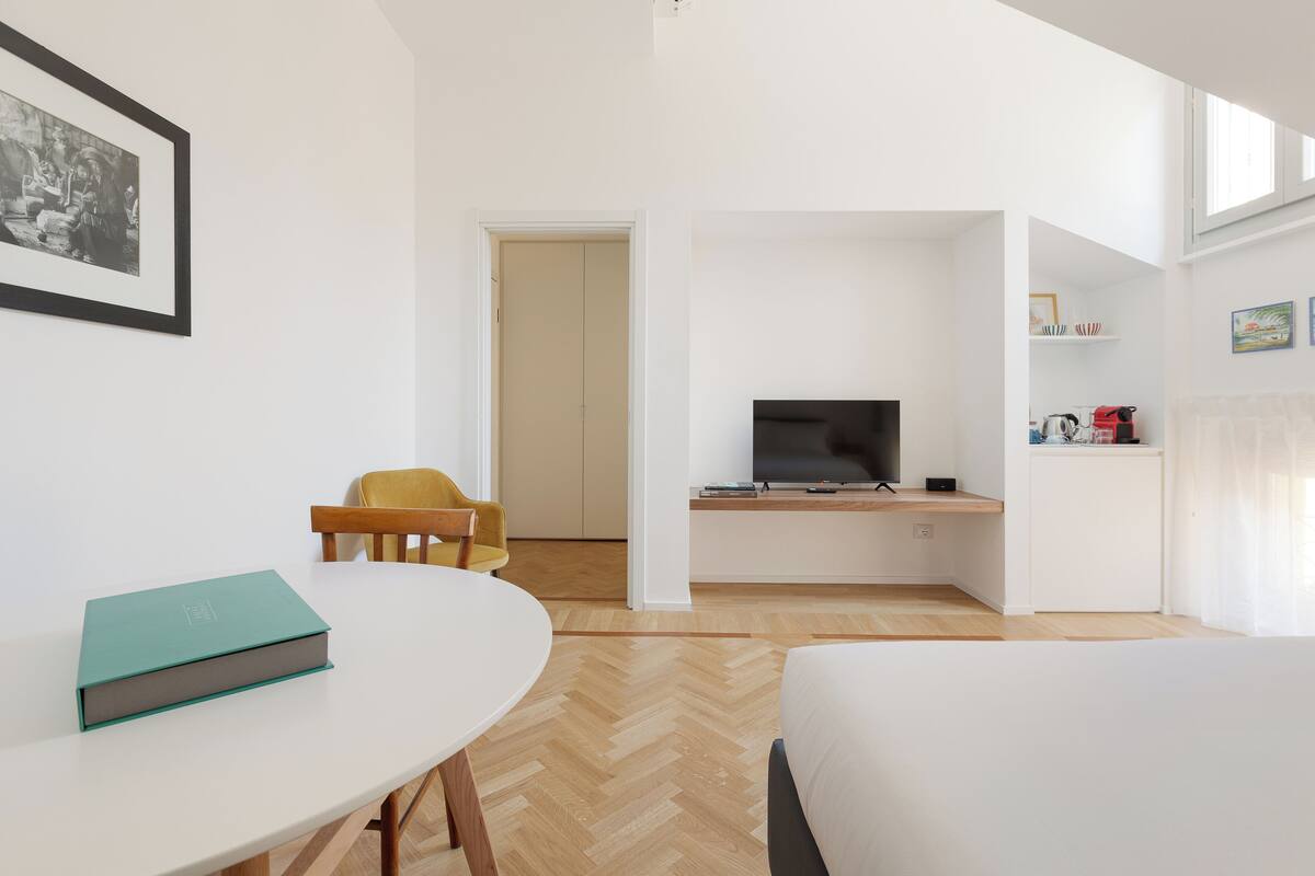 A modern hotel room with a bed, a desk, a chair, a television, and a bookshelf.